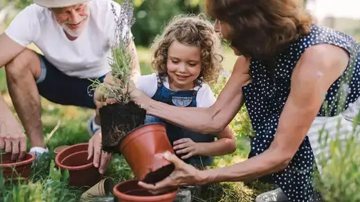 Young girl in denim overalls holds potted plant as woman assists outdoors