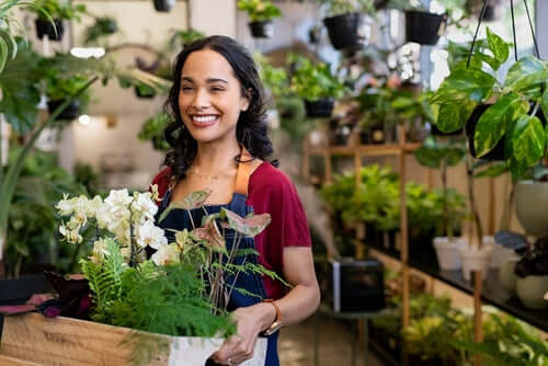 Smiling woman in red shirt and blue apron holds crate of lush green ferns and white flowers from garden shop