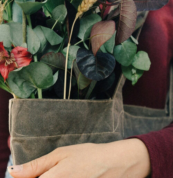 A dark olive waxed canvas apron with a sturdy, textured weave and visible stitching holds a vibrant bouquet of greenery and red flowers.