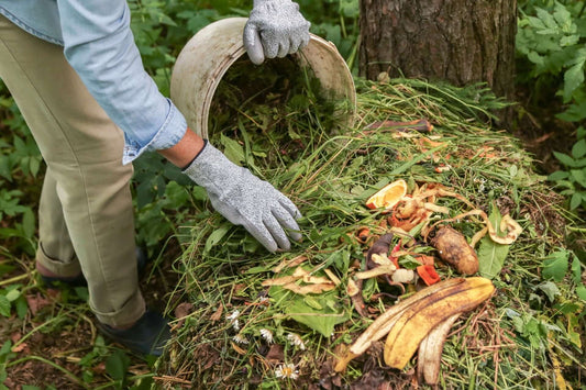 Person in gray gloves dumping garden waste from white bucket onto compost pile