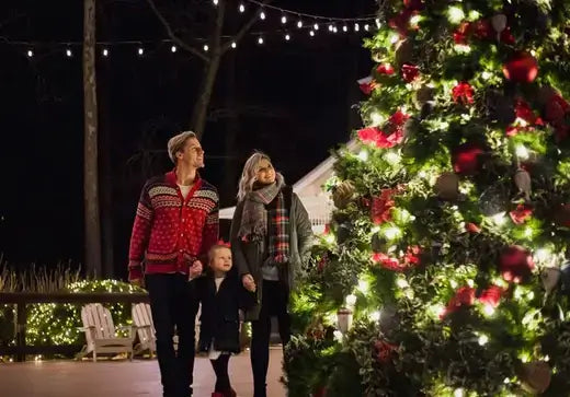 Family of three by brightly lit cedar Christmas tree with red ornaments